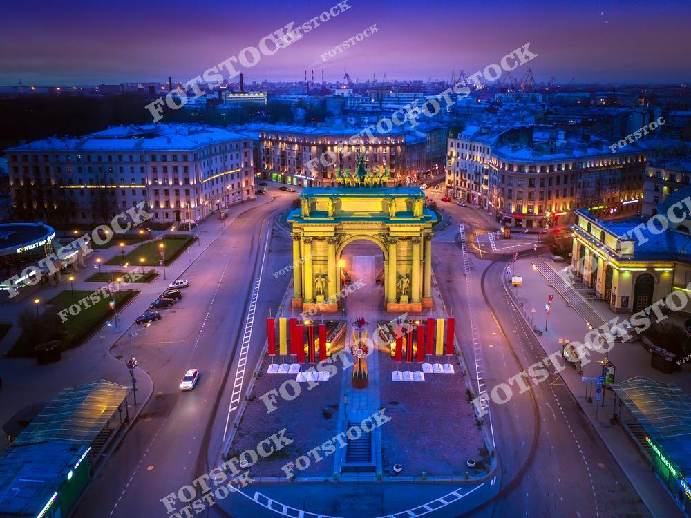 На фотографии запечатлены Нарвские триумфальные ворота, ярко освещённые ночными огнями. Это монумент в стиле ампир, построенный в честь победы в Отечественной войне 1812 года. Окружающая площадь и архитектура создают гармоничный ансамбль. Ночная подсветка подчёркивает величие ворот и добавляет глубину городскому пейзажу.